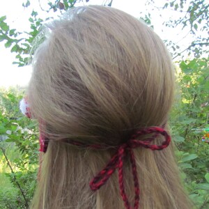 May include: A red and black crocheted headband with a white pom-pom on the side. The headband is tied in a bow at the back of the head.