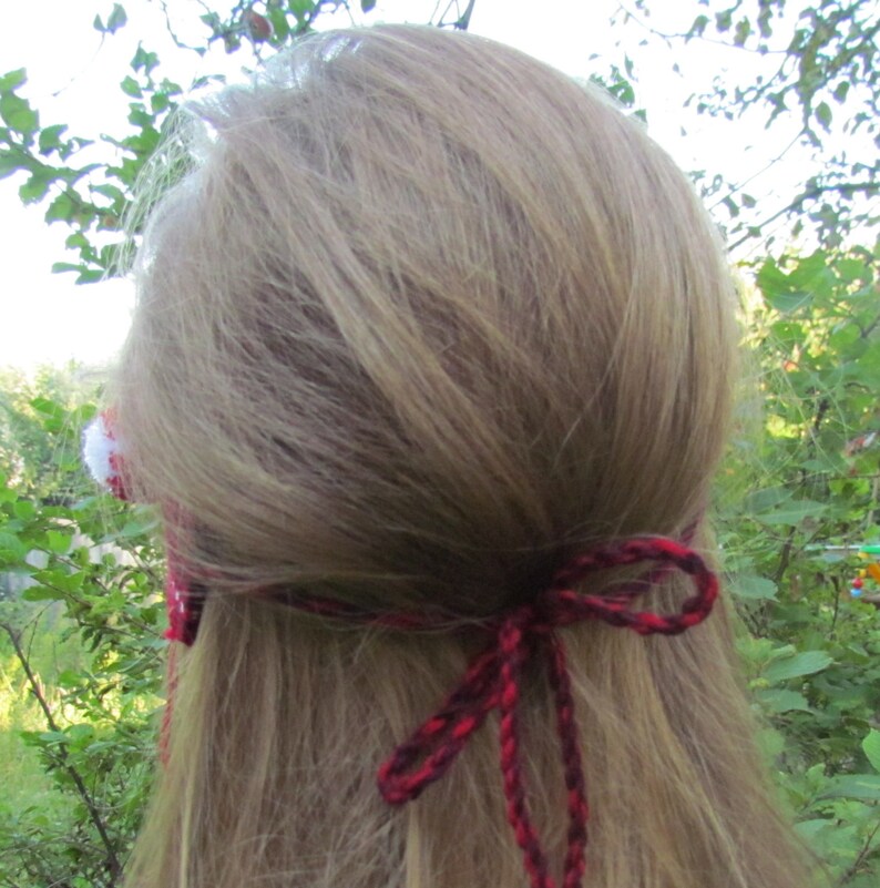 May include: A red and black crocheted headband with a white pom-pom on the side. The headband is tied in a bow at the back of the head.
