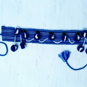 May include: A blue woven headband with white stripes and blue pom-poms. The headband has a white and blue design with small green accents.