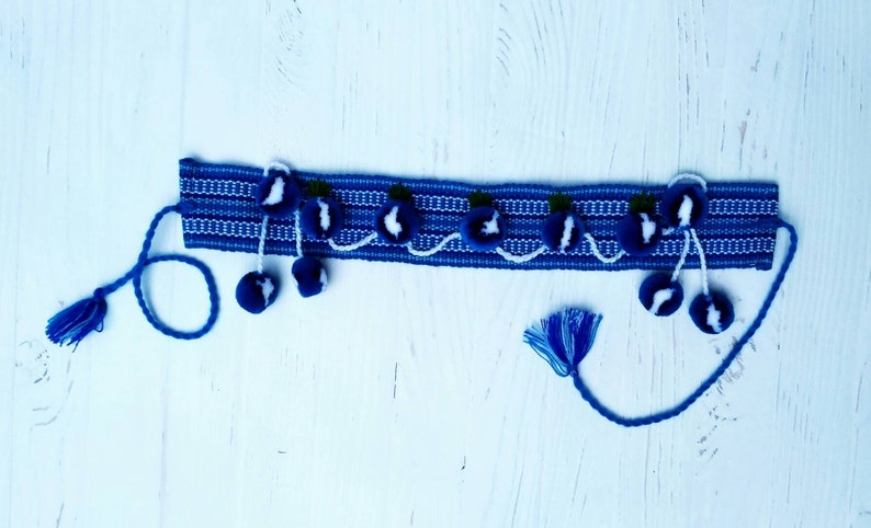 May include: A blue woven headband with white stripes and blue pom-poms. The headband has a white and blue design with small green accents.