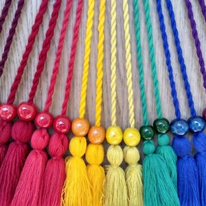 May include: A rainbow of colorful tassels with wooden beads. The tassels are made of yarn and are tied together with a string. The beads are round and have a smooth finish.