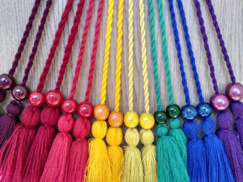 May include: A rainbow of colorful tassels with wooden beads. The tassels are made of yarn and are tied together with a string. The beads are round and have a smooth finish.