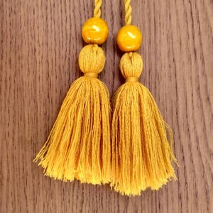 May include: Two mustard yellow tassels with round, mustard yellow beads at the top. The tassels are hanging against a brown wooden background.