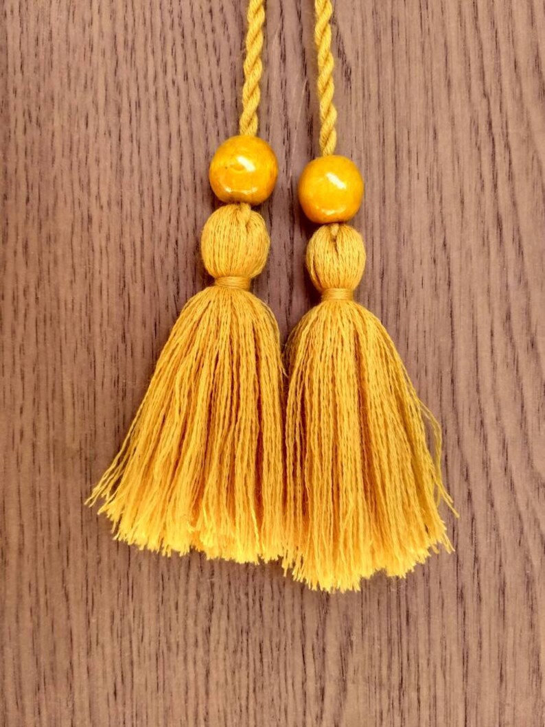 May include: Two mustard yellow tassels with round, mustard yellow beads at the top. The tassels are hanging against a brown wooden background.