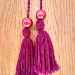 May include: Two burgundy tassels with red beads at the top. The tassels are hanging from a wooden surface.