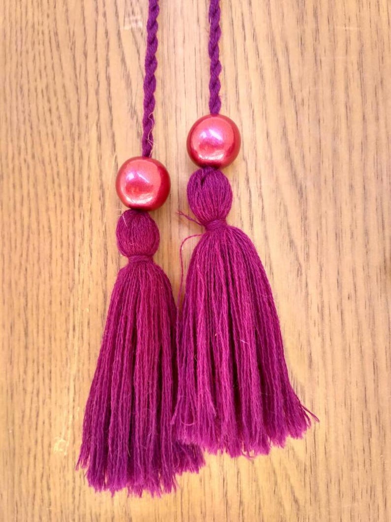 May include: Two burgundy tassels with red beads at the top. The tassels are hanging from a wooden surface.