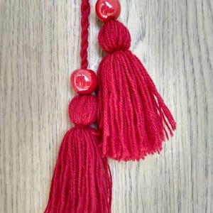May include: Two red yarn tassels with red bead accents hanging from a wooden surface.