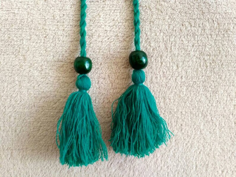 May include: Two green tassels with green beads at the top. The tassels are made of a soft, fuzzy material and are hanging from a white surface.
