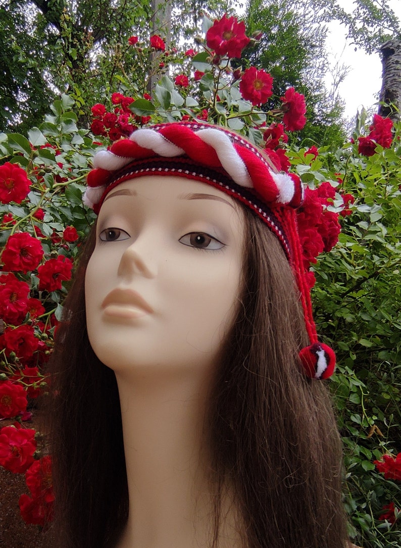 May include: A red and white braided headband with a white pom-pom on the end. The headband is worn by a mannequin with long brown hair.
