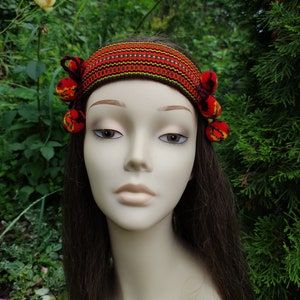 May include: A red, yellow, and green patterned headband with red pom-poms on each side. The headband is worn on a mannequin with long brown hair.