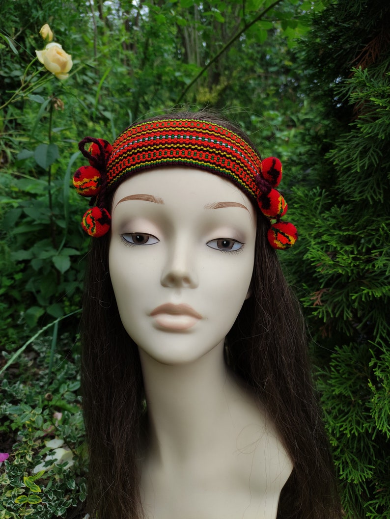 May include: A red, yellow, and green patterned headband with red pom-poms on each side. The headband is worn on a mannequin with long brown hair.