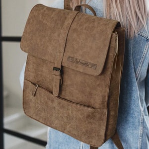 Könnte beinhalten: Ein brauner Rucksack aus Kunstleder mit einer schwarzen Schnalle und einer kleinen Tasche auf der Vorderseite. Der Rucksack hat das Wort "Autoinette" auf der Vorderseite geprägt.