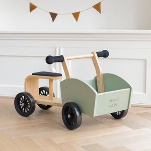 Könnte beinhalten: Ein hölzerner Spielzeugwagen mit einer grünen Aufbewahrungsbox und schwarzen Rädern. Der Wagen hat einen Holzgriff und einen schwarzen Sitz. Die Aufbewahrungsbox trägt den Namen "OSCAR".