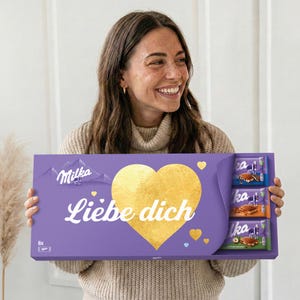Op de afbeelding: Een paarse geschenkdoos met Milka chocoladerepen, een gouden hart en de tekst "Liebe dich". De doos bevat zes chocoladerepen. Het Milka-logo staat linksboven.