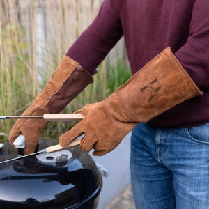 Op de afbeelding: Een persoon draagt ​​bruine leren grillhandschoenen met de tekst "Michael" en de initialen "LM" op de achterkant van de handschoenen. De handschoenen worden gebruikt om een ​​tang boven een zwarte grill te houden.