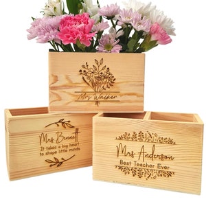 May include: Three wooden rectangular planters with engraved floral designs and names. One planter holds pink, white, and purple flowers. The text on the planters reads "Mrs Walker", "Mrs Bennett", and "Mrs Anderson Best Teacher Ever".