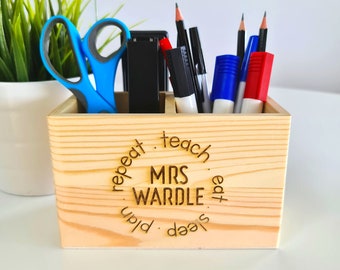 May include: Wooden desk organizer with four compartments. The organizer is engraved with the text "Sleep deep, plan, repeat, teach, eat, Mrs. Wardle". The organizer contains a pair of scissors, a black stapler, and several pens and markers.