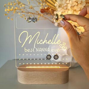 May include: A clear acrylic jewelry organizer with a light base. The organizer has a personalized message that reads "Michelle best NANNA ever" with two hearts. The organizer has a grid of holes for storing earrings and rings.