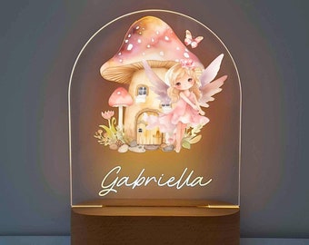 May include: A personalized night light featuring a fairy in a pink dress standing in front of a mushroom house. The light is made of acrylic and has a wooden base. The name "Gabriella" is written on the light.