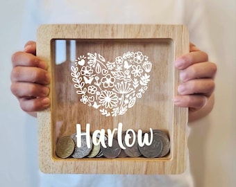 May include: A wooden coin bank with a clear front panel. The front panel features a white heart design with floral and butterfly details. The name "Harlow" is printed below the heart. Coins are visible inside the bank.