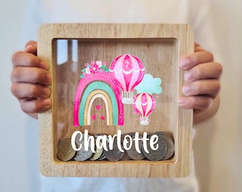 May include: Wooden piggy bank with a clear front panel. The front panel features a rainbow with hot air balloons and the name "Charlotte".