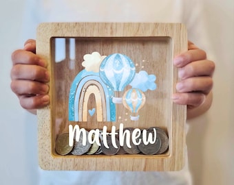 May include: A wooden shadow box with a clear front, filled with coins. The box features a colorful design of a rainbow, hot air balloons, and clouds in shades of blue, tan, and white. The name "Matthew" is printed in white across the bottom.