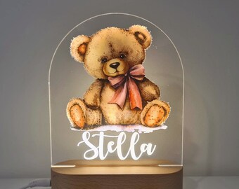 May include: A wooden night light with a clear acrylic dome featuring a watercolor illustration of a brown teddy bear wearing a pink bow. The name "Stella" is written in white on the base.