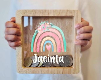 May include: A wooden piggy bank with a clear front panel. The front panel features a rainbow with a heart in the center and a floral design. The name "Jacinta" is printed below the rainbow. Coins are visible inside the piggy bank.
