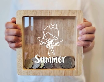 May include: A wooden coin bank with a clear front panel. The bank features a white outline of a fairy with wings and a bow. The name "Summer" is written below the fairy. The bank is filled with coins.