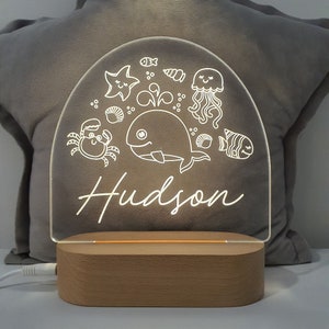 Puede incluir: Una luz nocturna de acrílico blanco con forma de semicírculo con una ballena, un cangrejo, una estrella de mar, una medusa y conchas. El nombre "Hudson" está escrito en cursiva debajo del diseño. La luz se asienta sobre una base de madera.
