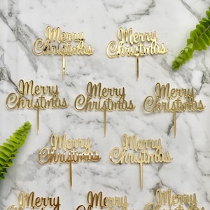 Pode incluir: Nove toppers de bolo dourados com a inscrição 'Feliz Natal' em letra cursiva. Os toppers estão sobre um fundo de mármore branco.