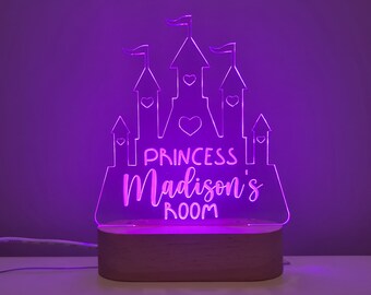 Puede incluir: Una luz nocturna LED de color morado con forma de castillo con el texto "Princess Madison's Room" en la parte delantera. La luz está sobre una base de madera.