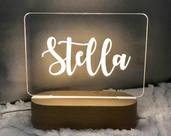 May include: A white acrylic sign with the name "Stella" written in a cursive font. The sign is illuminated from behind and sits on a wooden base.