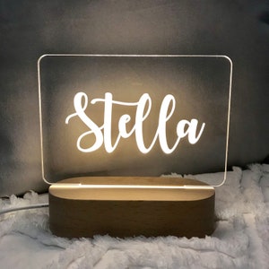 May include: A white acrylic sign with the name "Stella" written in a cursive font. The sign is illuminated from behind and sits on a wooden base.