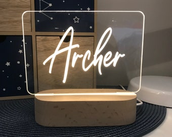May include: A white acrylic sign with the name "Archer" written in a cursive font, illuminated from behind by a warm white LED light. The sign is mounted on a wooden base.