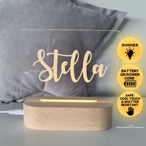 Op de afbeelding: Een gepersonaliseerd acryl nachtlampje met de naam "Stella" gegraveerd in gouden letters. Het lampje heeft een houten voet en een dimmer. Het kan worden gevoed door batterij of snoer. Het lampje is veilig, koel aan te raken en splintervrij.