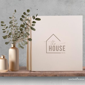 May include: A neutral-toned book with the text "The House & Co." is displayed on a wooden shelf. A gold vase with eucalyptus branches and a smaller gold vase are to the left of the book. The background is a gray concrete wall.