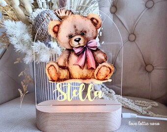 May include: A wooden night light with a clear acrylic panel featuring a watercolor illustration of a brown teddy bear wearing a pink bow. The name "Stella" is written in yellow script below the bear. The night light is plugged in and has a white cord.