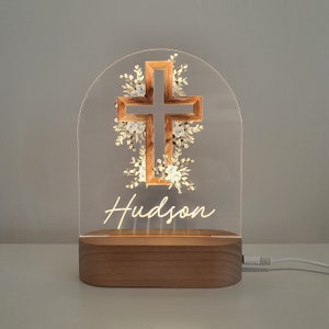 May include: A wooden base with a clear acrylic arch that features a brown cross with white floral accents. The name "Hudson" is written in white script below the cross. The light is on and illuminates the design.