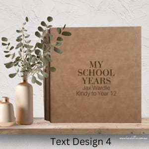 May include: A brown leather-bound photo album with the text "My School Years" and "Jax Wardle Kindy to Year 12" embossed on the front cover. The album is sitting on a wooden shelf with a vase of greenery in front of it.