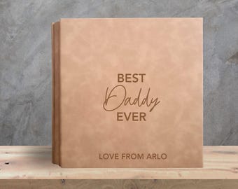 May include: A tan-colored book with the words "BEST Daddy EVER" engraved on the cover. Below the text is "LOVE FROM ARLO". The book is displayed on a wooden shelf against a gray background.