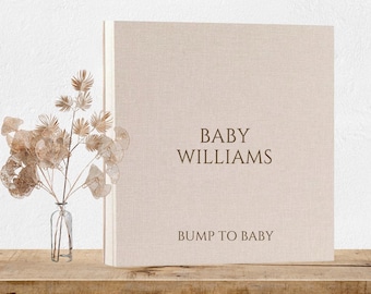 May include: A beige linen-covered baby memory book with the text "BABY WILLIAMS" and "BUMP TO BABY" in brown lettering. A small glass vase with dried flowers sits to the left of the book on a wooden shelf.