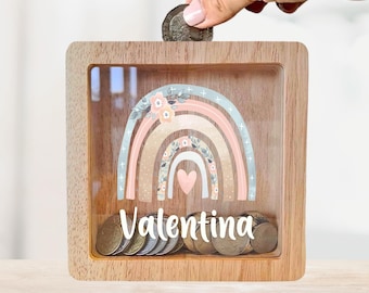 May include: A wooden rainbow piggy bank with a glass front. The rainbow is decorated with flowers and a heart. The name "Valentina" is written below the rainbow. Coins are visible inside the piggy bank.