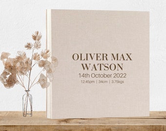 May include: A beige linen-covered photo album with the text "OLIVER MAX WATSON 14th October 2022 12:45pm | 34cm | 3.75kgs" printed in brown. The album is sitting on a wooden shelf with a vase of dried flowers in front of it.