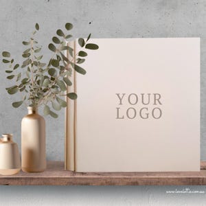May include: A neutral-toned book with the text "YOUR LOGO" is displayed on a wooden shelf. A gold vase with eucalyptus branches and a smaller gold vase are to the left. The background is a concrete wall.