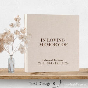 May include: A beige linen-covered photo album with the text "IN LOVING MEMORY OF" and "Edward Johnson 22.3.1944 - 15.1.2024" printed in brown. The album is on a wooden shelf with a vase of dried flowers in the foreground.