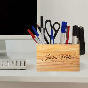 Puede incluir: Un organizador de escritorio de madera con seis compartimentos, que contiene varios artículos de oficina, incluidos bolígrafos, tijeras y una grapadora. El organizador está personalizado con el nombre "Jessica Miller" y el título "Office Manager".