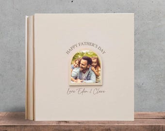 May include: A cream-colored photo album with the text "Happy Father's Day" above a family photo. The album also has the text "Love Eden & Clare" and "Text Design 3". The album is on a wooden shelf.