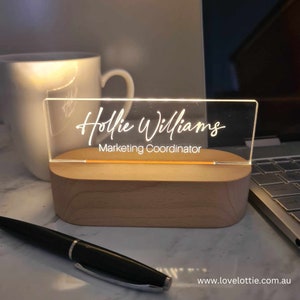 May include: A white acrylic desk sign with the name "Hollie Williams" and the title "Marketing Coordinator" in a cursive font. The sign is illuminated from behind and sits on a wooden base.