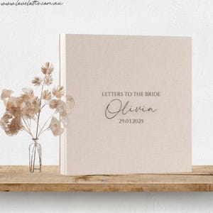 May include: A beige linen-covered book titled "Letters to the Bride" with the name "Olivia" and the date 29.03.2025 printed on the cover. A small glass vase with dried flowers sits to the left of the book on a wooden shelf.